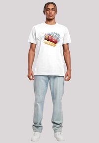 Un tricou alb din bumbac, cu un grafic colorat al lui Lightning McQueen, prezentând elemente de design în nuanțe de albastru și portocaliu, asortat cu blugi de un albastru deschis.