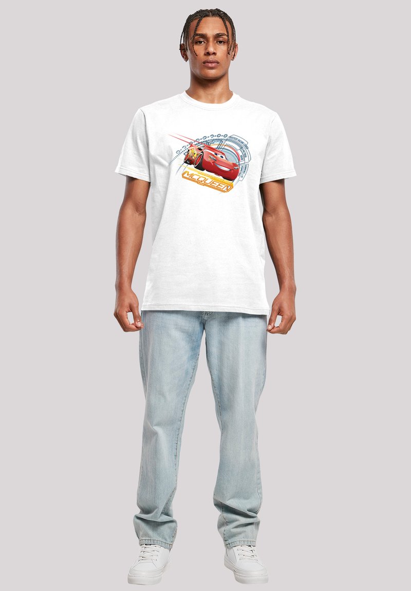 Un tricou alb din bumbac, cu un grafic colorat al lui Lightning McQueen, prezentând elemente de design în nuanțe de albastru și portocaliu, asortat cu blugi de un albastru deschis.