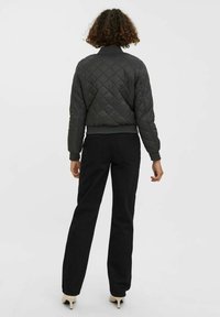 Czarna pikowana kurtka typu bomber z dopasowanym pasem, ściągaczami na rękawach i gładkim połyskiem, zestawiona z czarnymi spodniami i jasnym obuwiem.