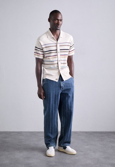 Gebreid shirt met korte mouwen in crème met multikleurige strepen, gecombineerd met donkerblauwe gestreepte pantalons en witte sneakers met een donkere accent.