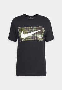 Camiseta de algodón negra con un gran Swoosh blanco de Nike sobre un patrón de camuflaje verde, mangas cortas y cuello redondo.