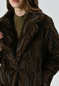 Cappotto in finta pelliccia marrone con una trama morbida, caratterizzato da un ampio colletto e un motivo a righe verticali. Accompagnato da un top in maglia verde sottostante.