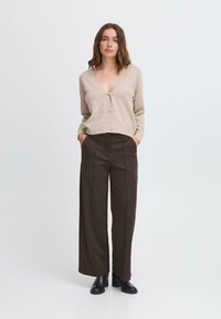 Cardigan en tricot beige avec fermeture à boutons, associé à un pantalon large marron taille haute et des bottines noires. Texture lisse, coupe décontractée.