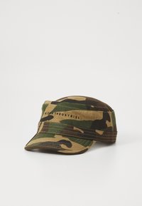 Gorra de estilo militar con patrón de camuflaje y visera corta, con el texto "Alpha Industries" en la banda frontal, descansando sobre una superficie blanca.