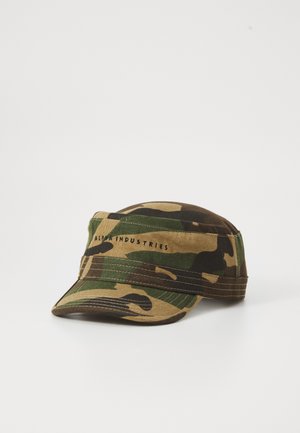 Hattu militaire avec un motif camouflage et une courte visière, portant le texte "Alpha Industries" sur la bande avant, reposant sur une surface blanche.