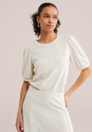 Femeie purtând un top crem cu mâneci scurte și textură, și o fustă cu talie înaltă asortată, cu model de frunze, pozând cu mâna pe șold, purtând cercei tip cerc.