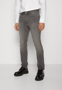 Man som har på sig skräddarsydda grå jeans, svarta snörskor och en vit långärmad skjorta med knappar, står mot en enfärgad vägg.