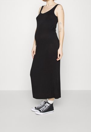 Personne enceinte portant une robe maxi noire sans manches et des baskets montantes noires, debout devant un fond blanc uni.