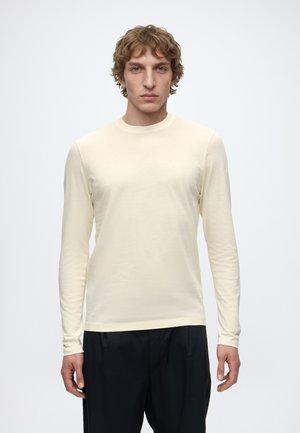 Junger Mann mit lockigem Haar, der ein cremefarbenes Langarmshirt und schwarze Hose trägt, steht vor einem schlichten weißen Hintergrund.