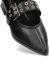 Zapato de tacón alto de cuero negro con punta afilada, que presenta una tira ancha adornada con ojales de plata y una hebilla para ajuste.