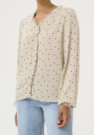 Femme portant un chemisier beige à manches longues avec des pois noirs, un devant à volants, et un jean bleu clair sur un fond uni.