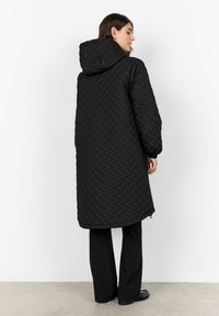 Palton negru matlasat cu glugă, cu un model țesut. Mâneci lungi și largi și tiv rotunjit. Modelul este prezentat cu pantaloni și pantofi negri.