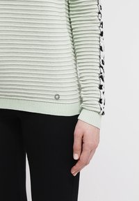 Hellgrüner, gerippter Pullover mit schwarzen Blumenakzenten entlang der Ärmel. Verfügt über eine strukturierte Oberfläche und ein kleines Logo-Detail am Saum.