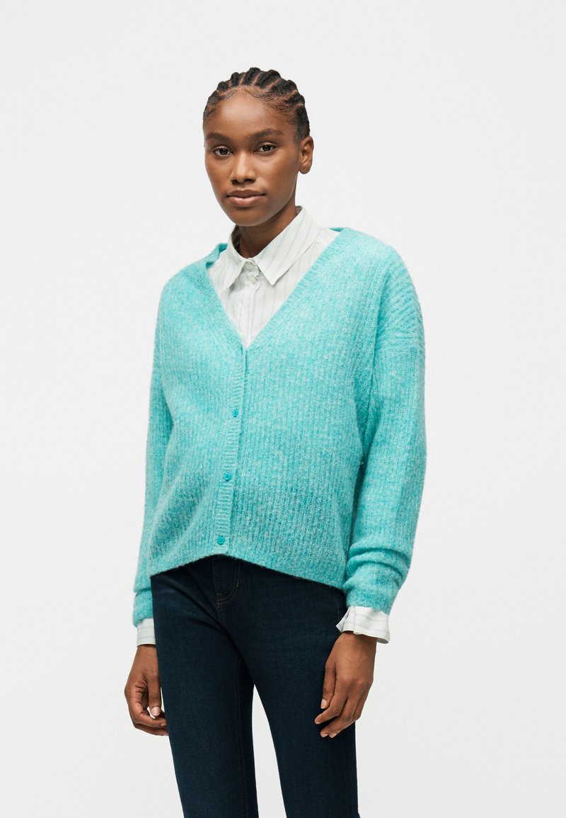 Γυναίκα με πλεξούδες, φορώντας μια τυρκουάζ ζακέτα με κουμπιά πάνω από ένα λευκό πουκάμισο με γιακά και σκούρο παντελόνι, στέκεται μπροστά σε ένα απλό φόντο.