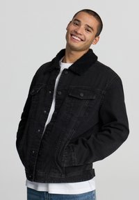 Giacca di jeans nera con un morbido colletto in pelliccia sintetica nera, chiusura con bottoni, due tasche sul petto e vestibilità comoda, indossata sopra una camicia bianca.