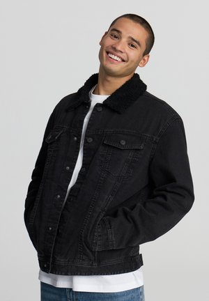 Giacca di jeans nera con un morbido colletto in pelliccia sintetica nera, chiusura con bottoni, due tasche sul petto e vestibilità comoda, indossata sopra una camicia bianca.