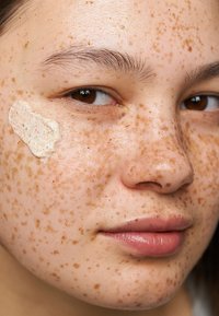 Ansiktsnära bild som visar hud med många fräknar och en krämisk exfoliant applicerad på kinden, vilket framhäver en texturerad, kornig yta.