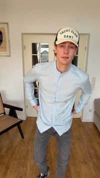 Noor mees, kes kannab heledat särki, halli teksapükse, tossusid ja "Yacht Club Gant" mütsi, seisab siseruumides käed puusas.