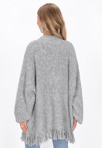 Grauer, übergroßer Strickpullover mit langen Ärmeln und fransigem Saum. Weiche Textur, lässiges Design. Über blauen Jeans getragen.