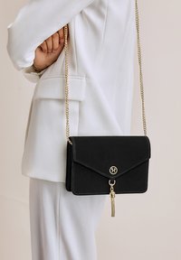 Borsa a tracolla in suede nero con tracolla in catena dorata, chiusura a patta magnetica, decorazione con logo e dettaglio con nappina sul davanti.