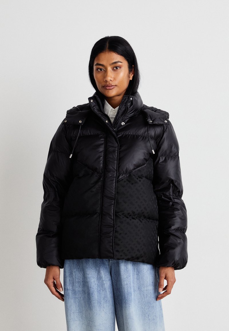 Mustapintainen pehmeä takki, jossa on kiiltävä pinta, korkealla kauluksella, nyöreillä ja toisella puolella geometrinen kuvio. Yhdistetty vaaleansinisiin farkkuihin.