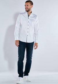 Weißes Button-Down-Hemd mit dunklen Knöpfen, kombiniert mit dunkelblauen Jeans und weißen Turnschuhen. Das Hemd hat lange Ärmel und ein schlankes Design.