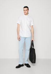 Fiatal férfi állva tart egy fekete bőrtáskát, fehér rövid ujjú gombos ingben, világoskék farmerral és fekete cipővel.