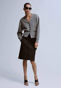 Šedý zapínací svetr, černá džínová sukně a černé nazouvací sandály. Modelka pózuje s rukama v kapsách před světlým pozadím.