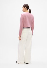 Pulover roz scurt cu mâneci lungi, dintr-un material texturat. Asociat cu pantaloni largi albi, cu buzunare la spate. Pantofi negri vizibili.