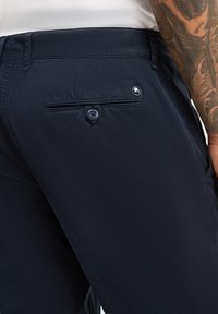Pantaloni blu navy con una trama liscia, dotati di tasca posteriore con chiusura a bottone e un piccolo dettaglio con logo.
