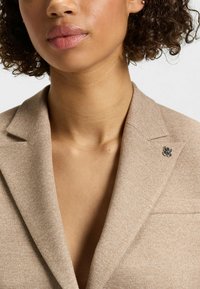 Blazer beige con un tessuto testurizzato, caratterizzato da un collo stretto e un logo del marchio argentato sul lato sinistro. Nessun motivo aggiuntivo visibile.