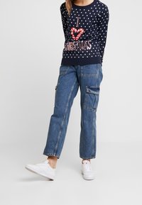 Enfant portant un pull bleu marine à pois avec le texte « J'aime Noël », un jean cargo bleu et des baskets blanches sur fond blanc.