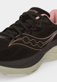 Gros plan d'une chaussure de course Saucony noire et rose avec des lacets noirs et une semelle crème montrant la texture en mesh et le logo sur le côté.