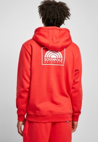 Red hoodie made of soft fabric, featuring a white Southpole logo with a rainbow design on the back. Includes a drawstring hood and ribbed cuffs.