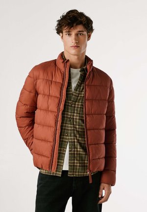 Chaqueta acolchada en naranja óxido, con patrón acolchado, cuello alto y cierre de cremallera, superpuesta sobre una camisa de cuadros verde con acentos marrones.