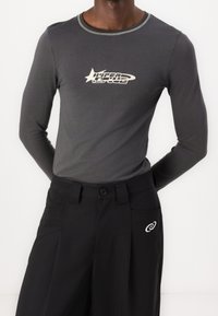 Muž oblečený v přiléhavém tmavě šedém triku s dlouhým rukávem a logem "HYPER SPEED" a černých kalhotách s malým bílým logem na kapse.