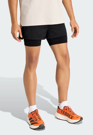 Czarne sportowe spodenki z wewnętrzną podszewką, połączone z jaskrawo pomarańczowymi butami do biegania z czarnymi akcentami i białymi skarpetkami.