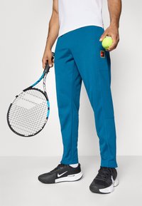 Vyras laiko teniso raketę ir kamuoliuką, vilkėdamas mėlynas Nike kelnes, juodus ir baltus Nike sportbačius bei baltą marškinėlį, stovintis ant vienspalvės fono.