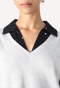 Top cinza com decote em V, apresentando uma gola preta com bolinhas brancas, destacando uma textura suave e um design em camadas.