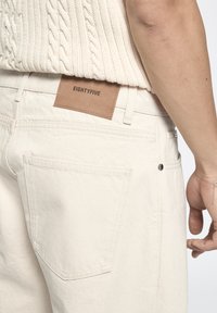 Helle beige Jeans mit glatter Textur, einem Lederbadge auf der Rückseite mit dem eingeprägten Schriftzug "EIGHTYFIVE" und Rückentaschen mit sorgfältigen Nähten.