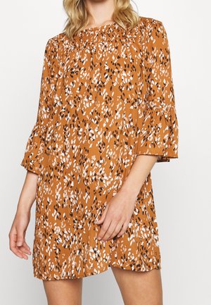 Frau trägt ein locker sitzendes, rost-orangefarbenes Kleid mit weißen und schwarzen abstrakten Flecken, mit 3/4 Glockenärmeln und einem gerafften Ausschnitt.