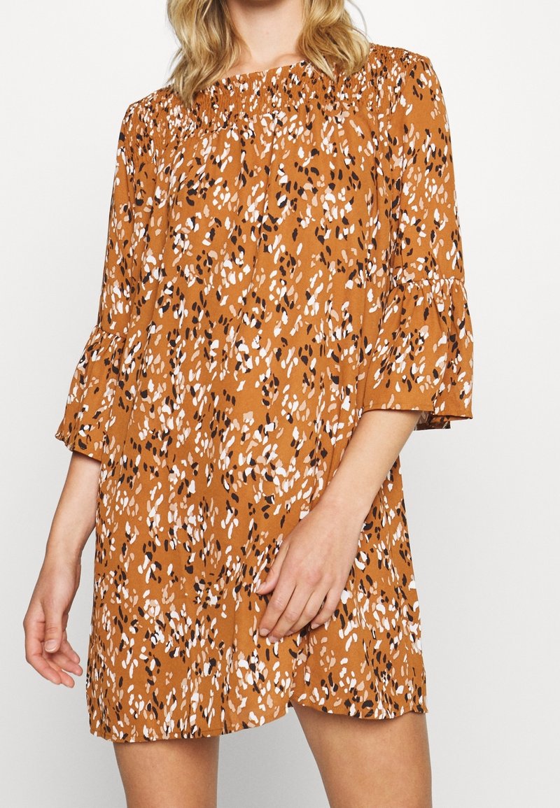 Femme portant une robe ample de couleur rouille avec des taches abstraites blanches et noires, dotée de manches cloche 3/4 et d'un col froncé.