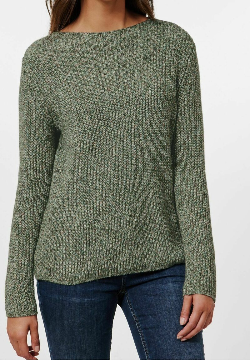 Femme portant un pull en tricot texturé vert et un jean bleu foncé, se tenant avec les bras détendus le long du corps contre un fond uni.