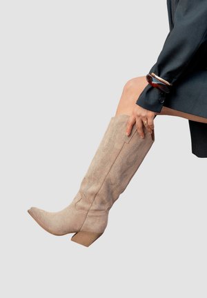 Femme portant des bottes de cowboy en suédine beige jusqu'au genou avec des détails brodés, un long manteau noir et des bracelets marron au poignet.
