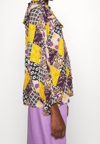 Blouse multicolore ornée de motifs floraux, à carreaux et de lèvres en jaune, violet et noir ; encolure à volants et manches longues.