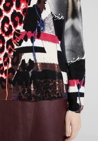 Pull tricoté présentant un mélange d'imprimés animaliers et de motifs audacieux en rouge, noir et blanc, avec une finition texturée et des accents bleus.