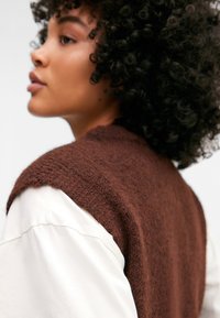 Brun fuzzy vest med en teksturert finish, båret over en kremfarget langermet skjorte. Vestet har rund hals og er ermeløs.