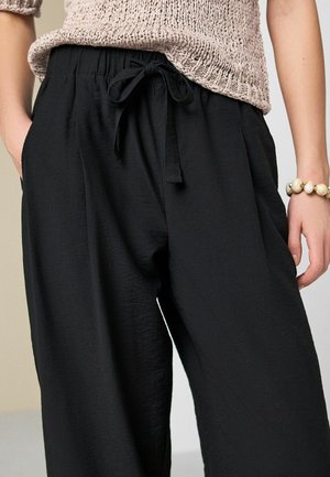 Lockere schwarze Kordelzug-Hose, getragen mit einem gestrickten beigen Top, Hände in den Taschen und ein Perlenarmband am rechten Handgelenk.