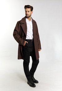Brauner Wollmantel mit großem Kragen, doppelter Knopfleiste und schwarzen Knöpfen. Über einem weißen Hemd getragen und kombiniert mit schmalen schwarzen Hosen und Stiefeln.