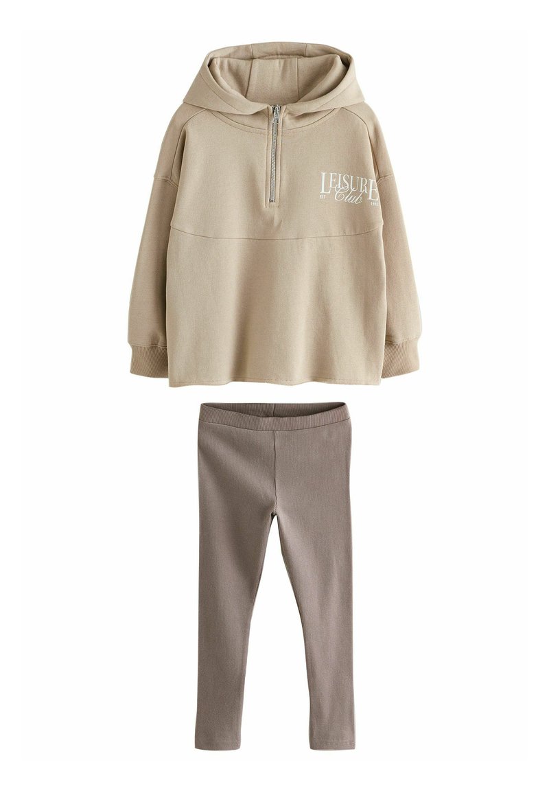 Beige hoodie med dragkedja och framtryckt lettering, kombinerad med ruggade bruna leggings. Mjuk bomullsmaterial, avslappnad passform och minimal design.
